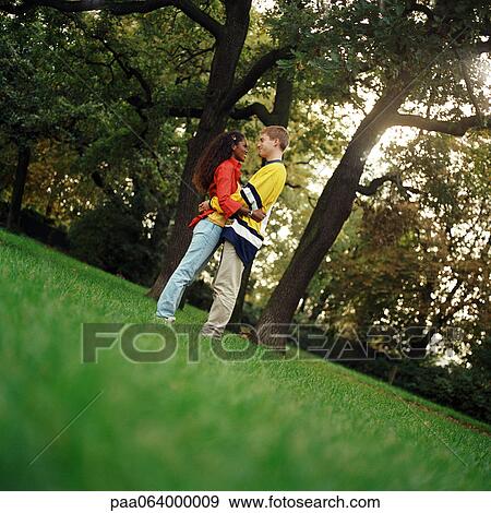 Stock Photo - Young man and woman standing on grass, embracing, side view, full length. Fotosearch
