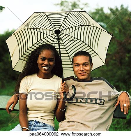 Stock Image - Young man and young woman smiling and sitting under umbrella outside. Fotosearch
