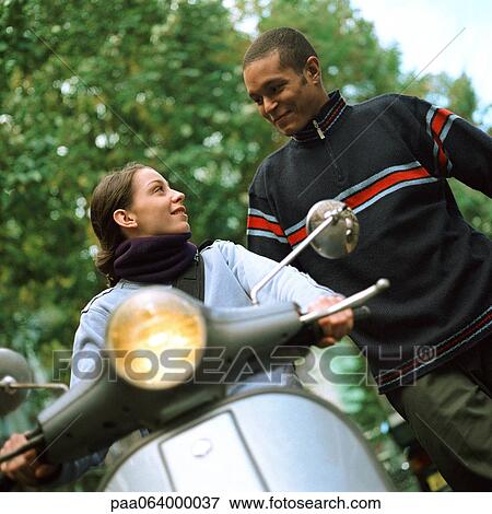 Stock Photo - Young man standing next to young woman on motor scooter, low angle view. Fotosearch