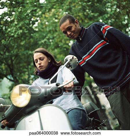 Stock Photo - Young man standing next to young woman on motor scooter. Fotosearch