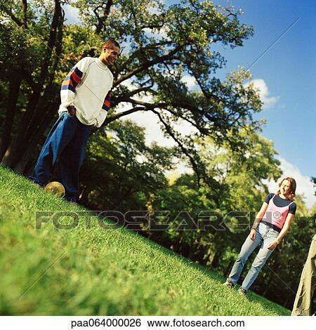 Stock Photograph - Young man standing on grass with soccer ball, young woman looking at him, low angle view. Fotosearch