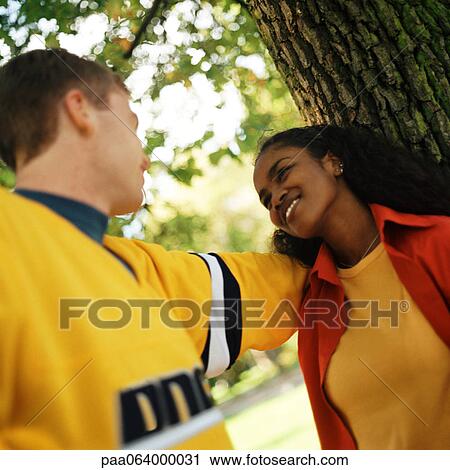 Young woman leaning against tree looking up at young man View Large Photo Image Stock Image - Young woman leaning against tree looking up at young man. Fotosearch