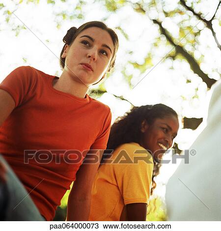 Stock Image - Young women sitting next to each other outside, low angle view. Fotosearch