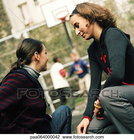Stock Image - Young women sitting together, young men in background playing basketball.. Fotosearch