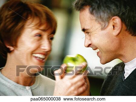 Stock Photo - Man and woman smiling at each other, holding apple between them, close up, blurred. Fotosearch
