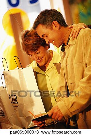 Stock Image - Man and woman with shopping bags, paying for purchase with check, waist up. Fotosearch