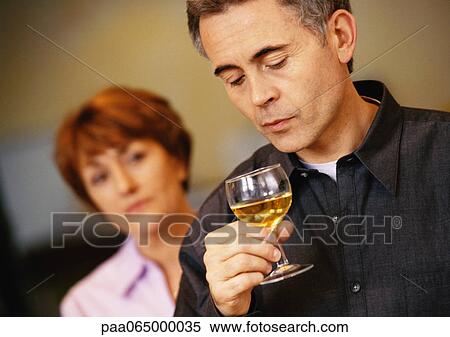 Stock Photography - Man holding and looking at wine glass, woman blurred in background, head and shoulders, close-up. Fotosearch