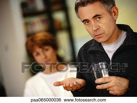 Stock Photograph - Man holding glass in one hand and pills in other, woman blurred in background, head and shoulders. Fotosearch