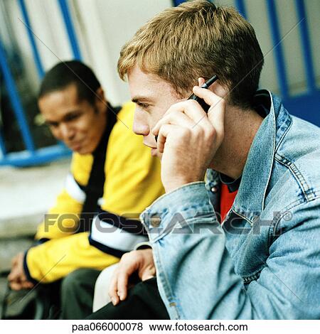 Stock Photo - Young men sitting together, close up of one on cell phone, side view. Fotosearch