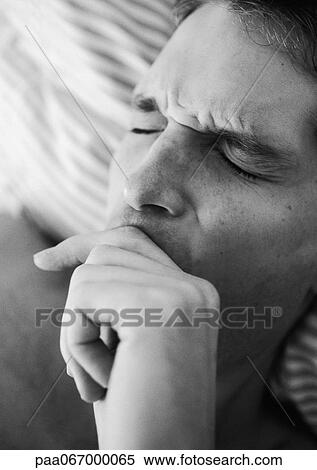 Stock Photography - Man's face with eyes closed and furrowed brow, hand over mouth, lying on bed, close-up, black and white.. Fotosearch