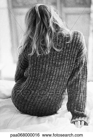 Stock Photograph - Woman sitting on bed in sweater, rear view, black and white.. Fotosearch