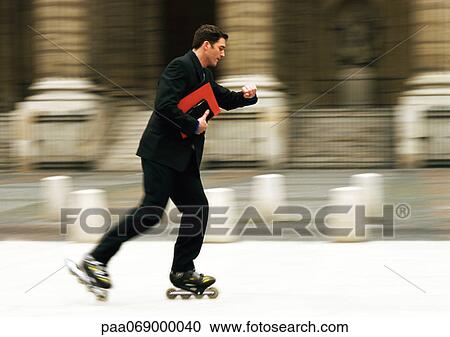 Stock Image - Businessman rollerblading in front of building and checking time, blurred.. Fotosearch