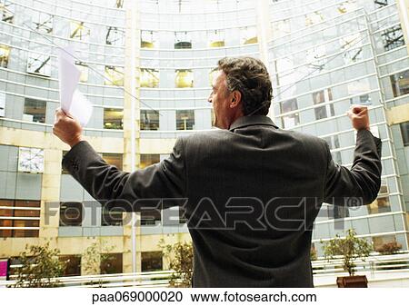 Stock Image - Businessman standing in front of building, arms raised in victorious gesture, rear view . Fotosearch