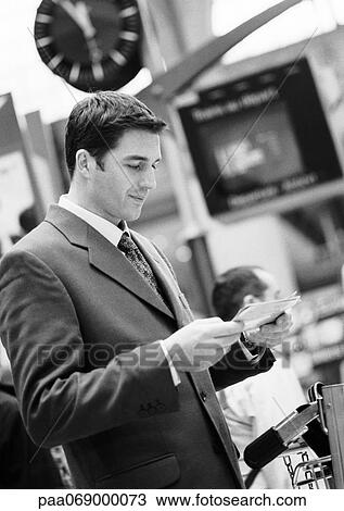 Stock Image - Businessman standing in terminal, looking at tickets, black and white.. Fotosearch