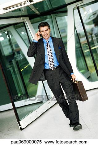 Stock Photography - Businessman using cell phone, walking away from glass doors, full length. Fotosearch