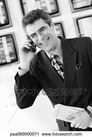 Stock Image - Businessman using cellular phone in airport, smiling and holding newspaper in front of monitors, black and white portrait.. Fotosearch