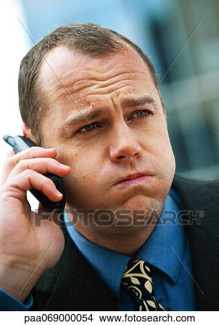 Picture - Businessman using cellular phone, puffing out cheeks and furrowing brow, portrait, close-up.. Fotosearch
