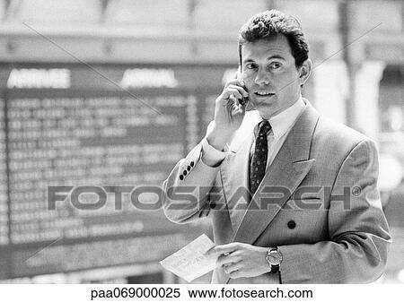 Stock Photography - Businessman using cellular phone, standing in front of arrival board, black and white.. Fotosearch