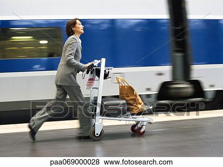 Stock Photo - Businesswoman rushing on train platform with cart and luggage, next to train, blurred.. Fotosearch