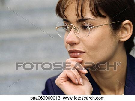 Stock Image - Dark-haired woman wearing glasses, holding chin with fingers, looking away, close-up.. Fotosearch