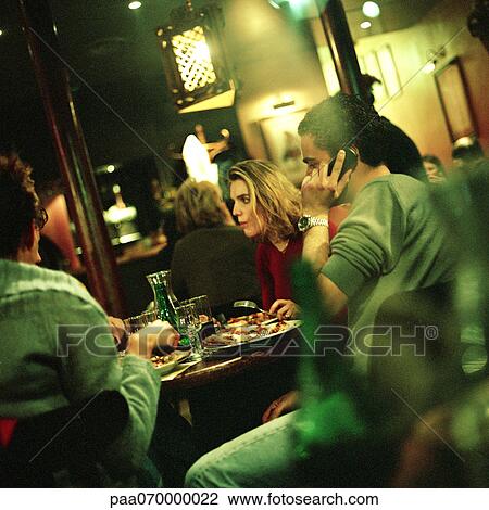 Stock Image - Group of young people eating at table in restaurant, one man using cell phone.. Fotosearch