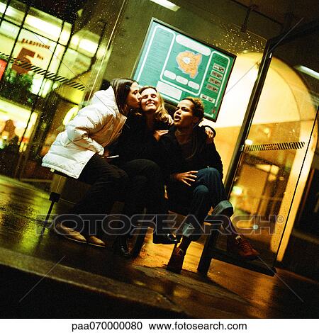 Stock Image - Three young women sitting in bus shelter at night. Fotosearch
