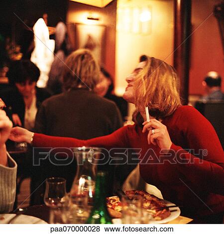 Woman sitting at table in restaurant, smoking a cigarette and reaching View Large Photo Image Stock Photo - Woman sitting at table in restaurant, smoking a cigarette and reaching. Fotosearch