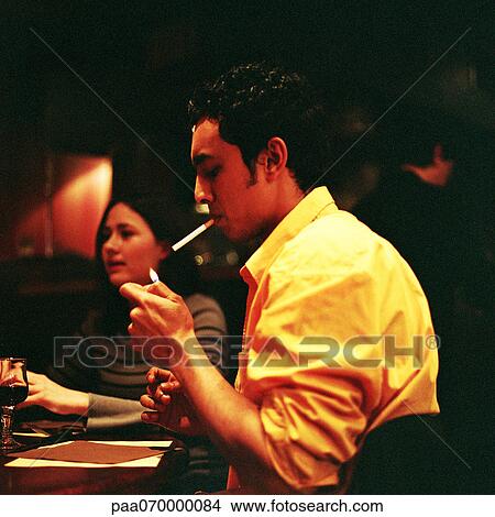 Young man lighting a cigarette in a cafe, young woman in background View Large Photo Image Picture - Young man lighting a cigarette in a cafe, young woman in background. Fotosearch