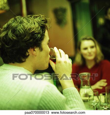 Stock Image - Young people sitting at table, young man in foreground drinking, rear view. Fotosearch