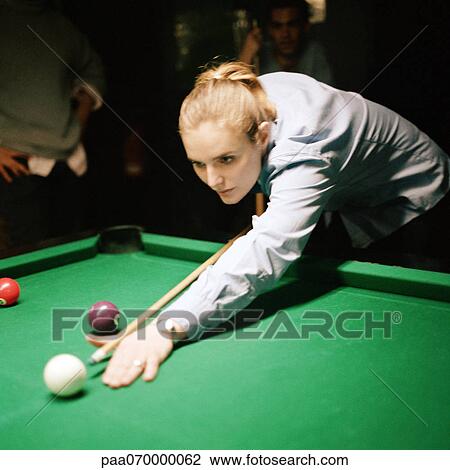 Stock Image - Young woman shooting pool, people standing in background. Fotosearch