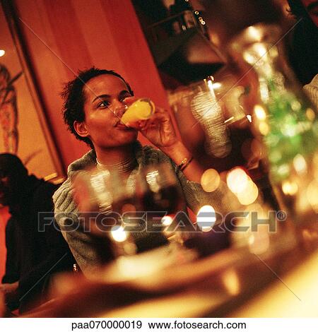 Young woman sitting at table, drinking, glasses blurred in foreground View Large Photo Image Stock Photo - Young woman sitting at table, drinking, glasses blurred in foreground. Fotosearch