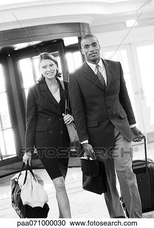 Stock Image - Businessman and businesswoman walking together inside building with luggage, b&w.. Fotosearch