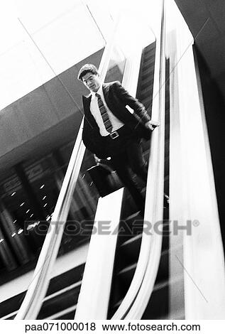 Stock Photo - Businessman going down escalator, looking down, b&w.. Fotosearch