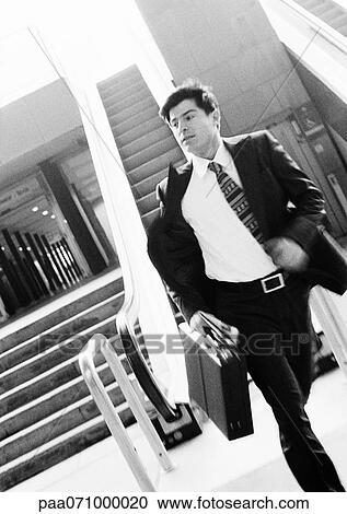 Stock Image - Businessman hurrying away from escalator, holding briefcase, blurred motion, b&w.. Fotosearch