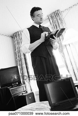 Businessman standing looking at agenda in hotel room, open laptop on bed in foreground, b&w. View Large Photo Image Stock Photo - Businessman standing looking at agenda in hotel room, open laptop on bed in foreground, b&w.. Fotosearch