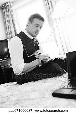 Stock Photo - Businessman working, sitting on edge of bed in hotel room, blurred, b&w.. Fotosearch