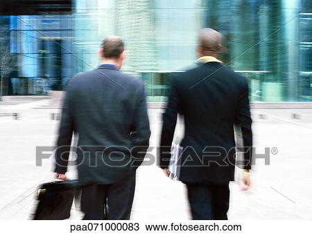 Stock Image - Businessmen walking towards building, rear view, blurred.. Fotosearch