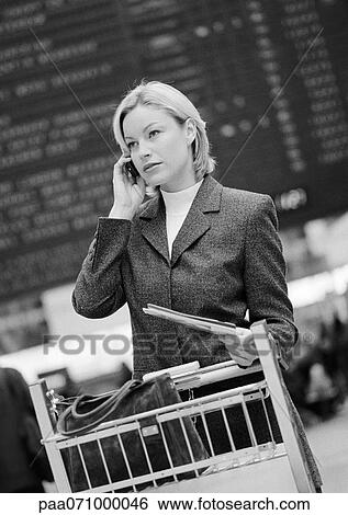 Stock Photograph - Businesswoman on cell phone with luggage caddy, in front of arrival and departure board, b&w.. Fotosearch