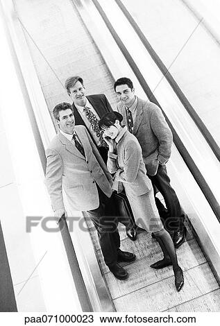 Stock Image - Group of business people on moving walkway smiling at camera, business woman in front on cell phone, b&w.. Fotosearch