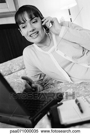 Stock Photography - Woman lying on bed, talking on cell phone and pointing at her laptop computer, b&w.. Fotosearch