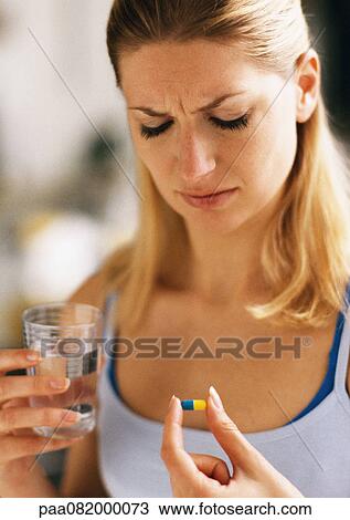 Stock Image - Woman holding pill and glass, looking down, head and shoulders, close-up. Fotosearch