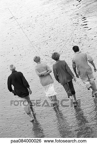 Stock Photograph - Four mature adults walking down the beach, view from rear, B&W.. Fotosearch