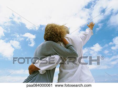 Stock Image - Mature couple embracing and looking up at the sky, rear view, low angle view. Fotosearch