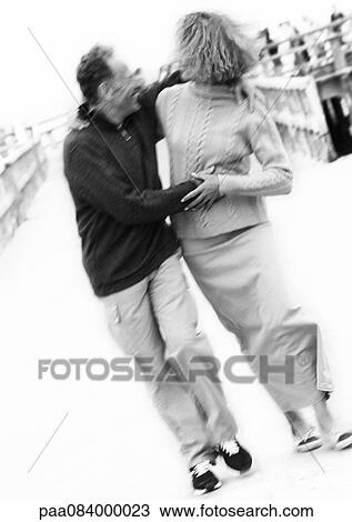 Stock Image - Mature couple holding each other on boardwalk, blurred, B&W.. Fotosearch