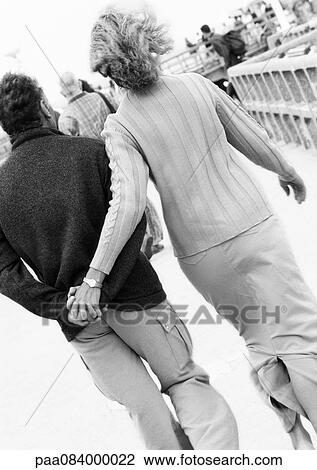 Stock Image - Mature couple walking down boardwalk holding hands, rear view, B&W.. Fotosearch