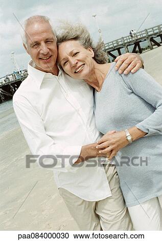 Stock Image - Mature couple with arms around each other on the beach, smiling at camera.. Fotosearch