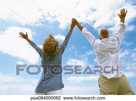 Stock Image - Mature couple with arms up in the air, holding hands, rear view, low angle view. Fotosearch
