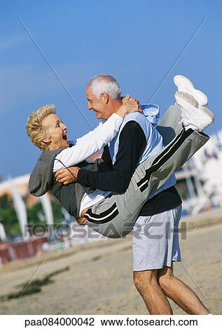 Stock Image - Mature man holding mature woman on beach. Fotosearch
