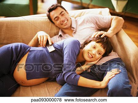 Stock Photo - Man and pregnant woman on sofa, woman lying on man's lap, man holding remote control, portrait. Fotosearch