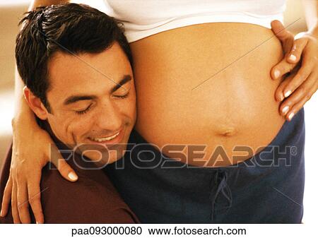 Stock Image - Man pressing cheek to pregnant woman's stomach, close-up. Fotosearch